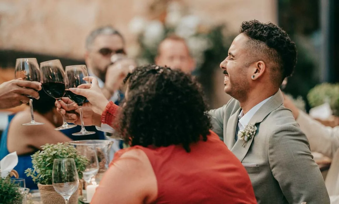 Eine Gruppe von Menschen sitzt an einem Tisch, lächelt und erhebt Weingläser zum Toast. Aufgenommen von Hochzeitsfotograf Mallorca Lukas P. Schmidt, zeigt die festliche Szene formelle Kleidung, üppige Pflanzen und sanfte Beleuchtung, die die Feier unterstreicht.