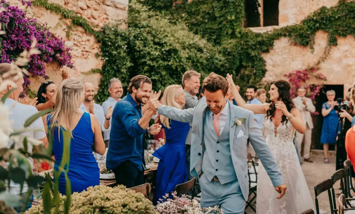Ein Bräutigam in einem hellblauen Anzug gibt den Hochzeitsgästen enthusiastisch ein High-Five, während er einen Gang im Freien hinunterläuft, gefolgt von seiner lächelnden Braut, während Lukas P. Schmidt, Hochzeitsfotograf Mallorca, den Jubel inmitten von üppigem Grün und Blumen einfängt.