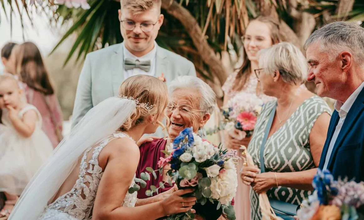 Eine Braut in weißem Kleid und Schleier umarmt und lächelt mit einer älteren Frau, die einen Blumenstrauß hält, umgeben von fröhlichen Gästen bei einer Hochzeit im Freien, aufgenommen von Hochzeitsfotograf Mallorca Lukas P. Schmidt.