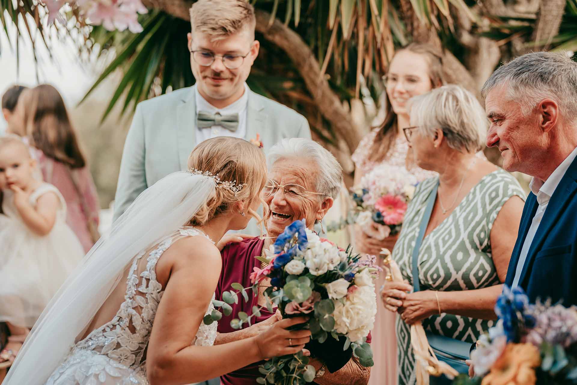 hochzeit mallorca 303 Eine Braut in weißem Kleid und Schleier umarmt und lächelt mit einer älteren Frau, die einen Blumenstrauß hält, umgeben von fröhlichen Gästen bei einer Hochzeit im Freien, aufgenommen von Hochzeitsfotograf Mallorca Lukas P. Schmidt.
