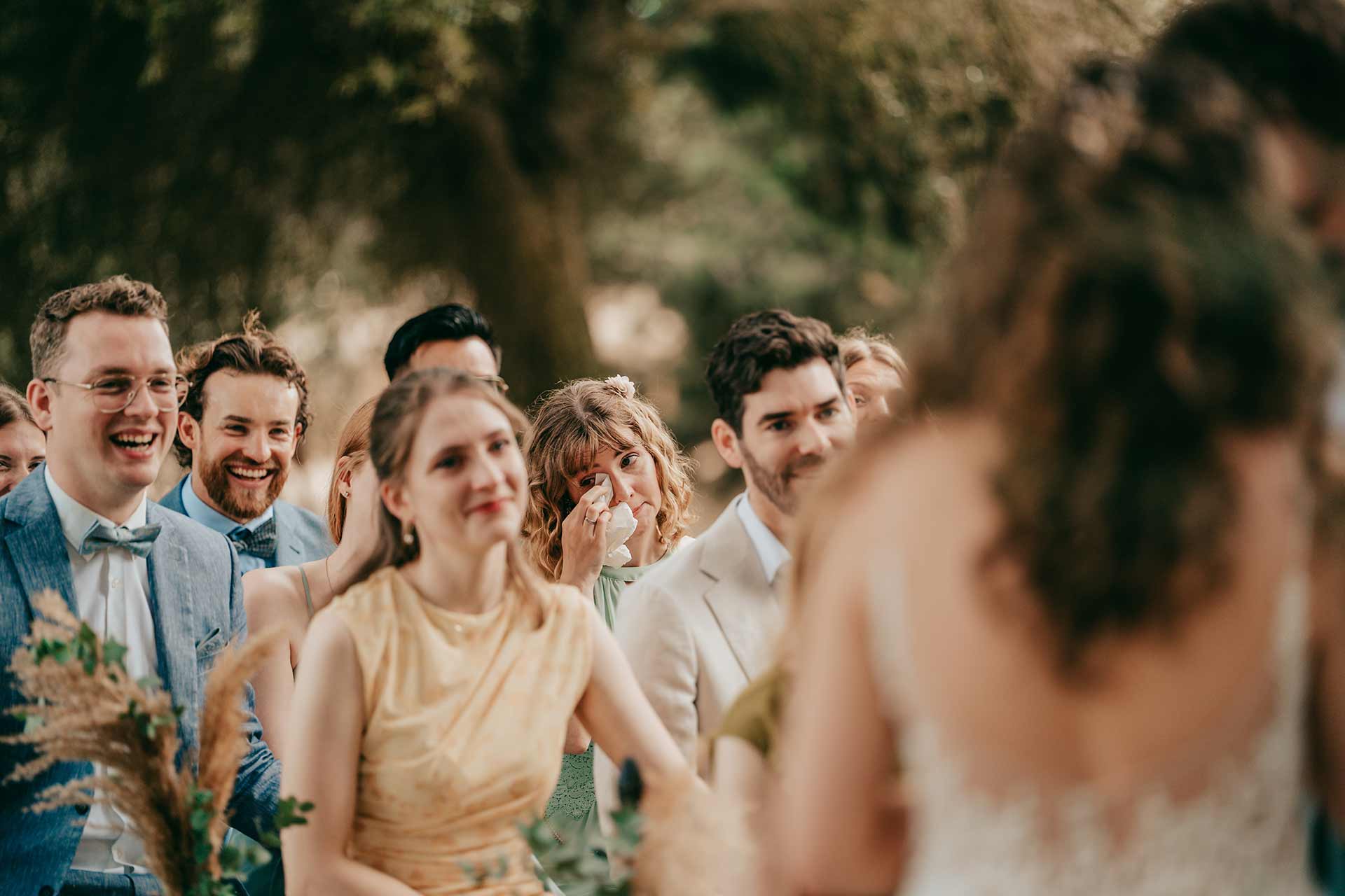 mallorca hochzeit 208 Eine Gruppe von Hochzeitsgästen sitzt im Freien, lächelnd und gerührt; eine Frau in einem grünen Kleid wischt sich die Tränen weg, während sie dem Paar, das teilweise von hinten zu sehen ist, während der Zeremonie zugewandt ist, aufgenommen von Hochzeitsfotograf Mallorca Lukas P. Schmidt.