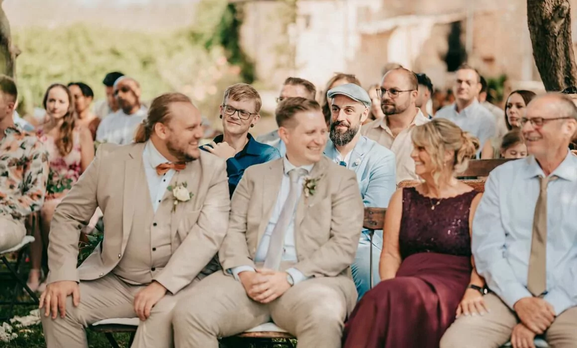 Eine Gruppe von formell gekleideten Menschen sitzt auf Bänken im Freien, wo eine Hochzeitszeremonie stattzufinden scheint. Hochzeitsfotograf Mallorca Lukas P. Schmidt fängt die fröhliche Stimmung ein, als die Gäste lächeln und sich unter natürlichem Sonnenlicht unterhalten.