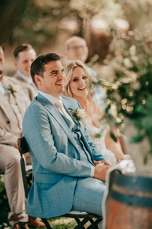 Eine Braut und ein Bräutigam sitzen bei einer Hochzeitszeremonie auf Finca Alaiar im Freien lächelnd und lachend nebeneinander. Der Bräutigam trägt einen hellblauen Anzug und die Braut ein weißes Kleid. Die Gäste sitzen hinter ihnen und die Szene ist von Grün umgeben.
